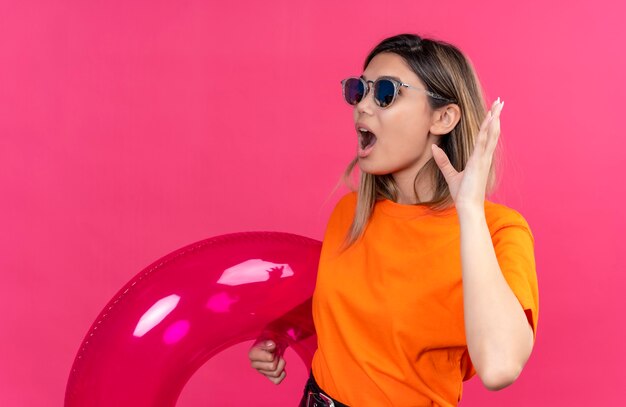 Eine überraschte hübsche junge Frau in einem orangefarbenen T-Shirt, das eine Sonnenbrille trägt, die Hand mit offenem Mund hebt, während rosa aufblasbaren Ring an einer rosa Wand hält