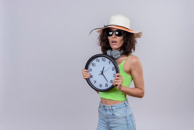 Eine überraschte junge Frau mit kurzen Haaren im grünen Erntedach, die Sonnenbrille und Sonnenhut hält, die eine Wanduhr auf einem weißen Hintergrund halten