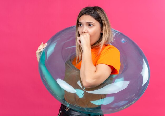 Eine überraschte reizende junge Frau in einem orange T-Shirt, das Seite mit Hand auf Mund schaut, während auf aufblasbarem Ring auf einer rosa Wand steht