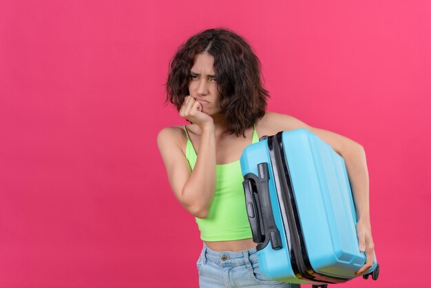 Eine verärgerte attraktive Frau mit kurzen Haaren, die grünes Erntedach hält und blauen Koffer hält, der wütend schaut