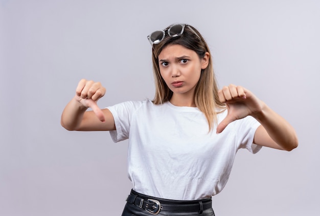 Eine verärgerte junge Frau im weißen T-Shirt im Sonnenbrillenlächeln, während Daumen nach unten zeigend