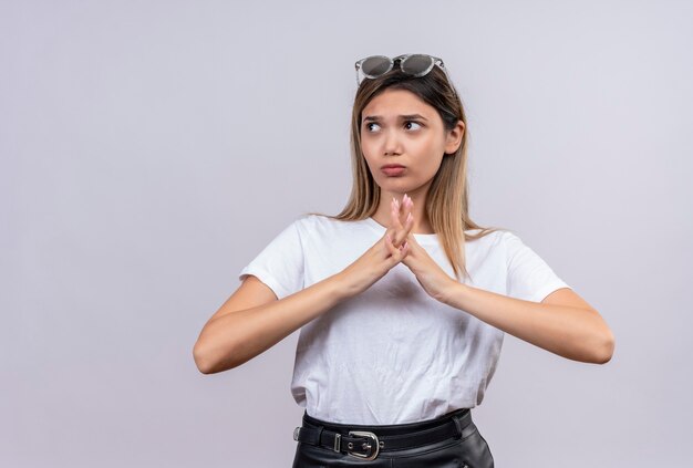 Eine verwirrte junge Frau im weißen T-Shirt in der Sonnenbrille, die Finger mit nachdenklichem Ausdruck auf einer weißen Wand faltet