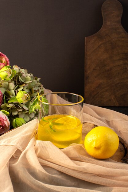 Eine Vorderansicht Gläser mit Saft Zitronensaft in transparenten Gläsern entlang der ganzen Zitrone und Blumen auf dem braunen Holzschreibtisch und dem grauen Hintergrund Cocktail Zitronengetränk