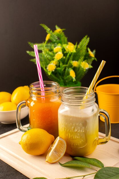 Eine Vorderansicht kalte Cocktails gefärbt in Glasdosen mit bunten Strohhalmen Zitronengrün lässt Blumen auf dem hölzernen cremefarbenen Schreibtisch und dunkel