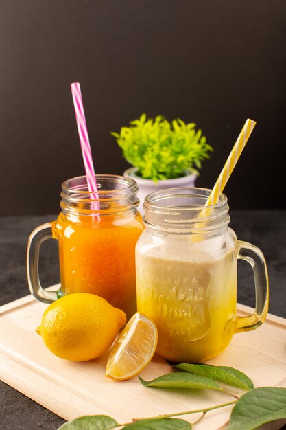 Eine Vorderansicht kalte Cocktails gefärbt in Glasdosen mit bunten Strohhalmen zitronengrünen Blättern auf dem hölzernen cremefarbenen Schreibtisch und dunkel