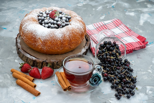 Eine Vorderansicht Obstkuchen mit frischem Blau, Beeren und zusammen mit einer Tasse Tee auf hellem, süßem Kuchenkekszucker