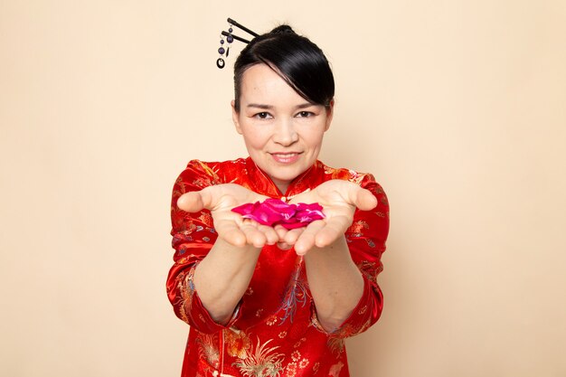 Eine Vorderansicht schöne japanische Geisha im traditionellen roten japanischen Kleid mit Haarstäbchen, die das elegante Lächeln der roten Rosenblätter auf der cremefarbenen Hintergrundzeremonie Japan Ost posierend halten