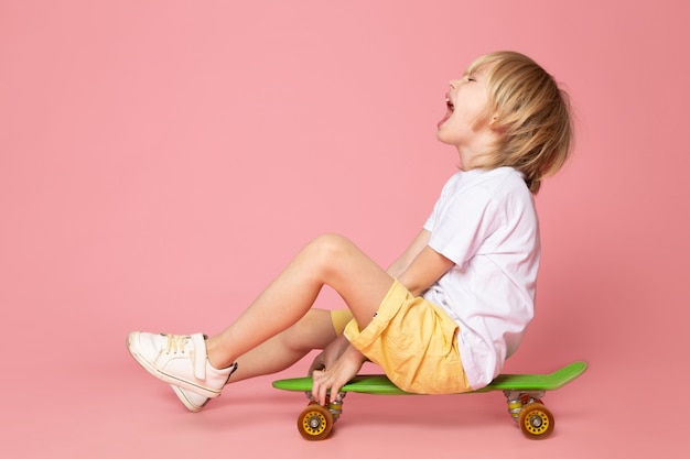 Eine Vorderansicht schreiender Junge blondes entzückendes niedliches grünes reitendes Skateboard auf dem rosa Raum