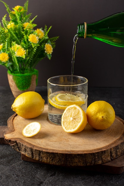 Eine Vorderansicht Wasser mit zitronenfrischem kühlem Getränk, das mit geschnittenen Zitronen zusammen mit ganzen Zitronen und Blumen im Dunkeln in das Glas gießt