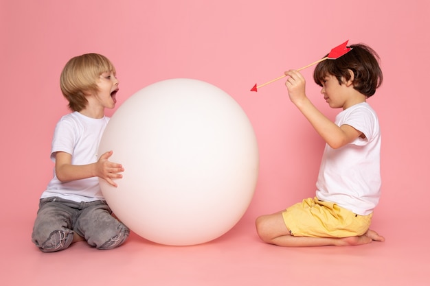 Eine Vorderansicht zweier Jungen, die durch weißen runden Ball in weißen T-Shirts auf dem rosa Boden miteinander spielen