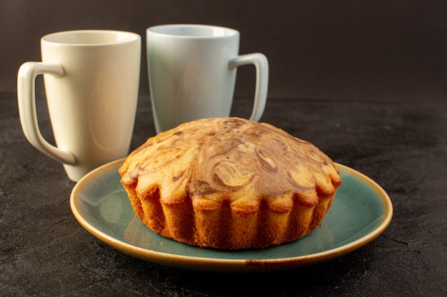 Eine vordere geschlossene Ansicht runder süßer Kuchen köstlicher leckerer Schokoladenkuchen innerhalb des blauen Tellers zusammen mit zwei weißen Tassen auf der Dunkelheit