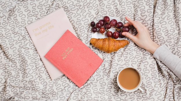 Eine weibliche Hand hält rote Trauben mit Hörnchen; Teetasse und Bücher auf Tischdecke