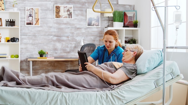 Eine weibliche Pflegekraft hilft einer alten behinderten Frau, die im Krankenhausbett liegt, einen digitalen Tablet-PC zu benutzen. Helles Zimmer mit großen Fenstern