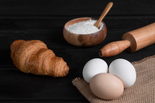 Einfache Croissants, Mehlschale und rohe Eier auf dunkler Holzoberfläche. Hochwertiges Foto