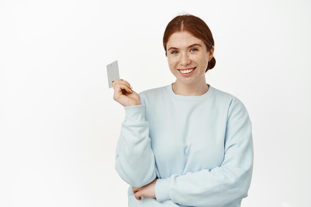 Einfache Zahlung. Lächelndes Ingwermädchen sieht selbstbewusst aus, hält eine Kreditrabattkarte in der Hand und sieht glücklich aus, geht weiter einkaufen, zahlt kontaktlos, weißer Hintergrund.