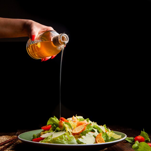 Einige Frau, die Öl auf köstlichen Salat in einem Teller auf hölzernem und schwarzem Hintergrund, Seitenansicht gießt. Platz für Text