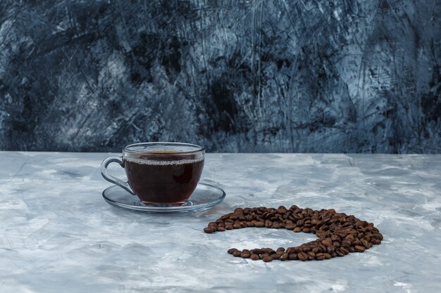 Einige Kaffeebohnen mit Tasse Kaffee auf dunkel- und hellblauem Marmorhintergrund, Nahaufnahme.