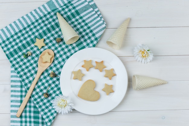 Einige Kekse mit Waffeltüten, Blumen im Teller und Holzlöffel auf Holz- und Küchentuchhintergrund, flach liegen.