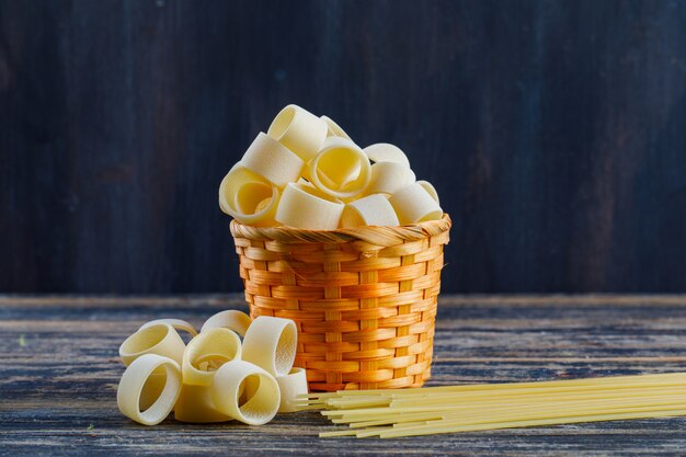 Einige Makkaroni mit Nudeln und Spaghetti in einem Eimer auf dunklem hölzernem Hintergrund, Seitenansicht. Platz für Text