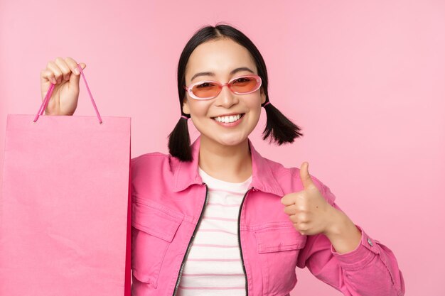 Einkaufen Stilvolles asiatisches Mädchen mit Sonnenbrille, das Tasche aus dem Geschäft zeigt und lächelnd Verkaufspromo im Geschäft empfiehlt, das über rosafarbenem Hintergrund steht