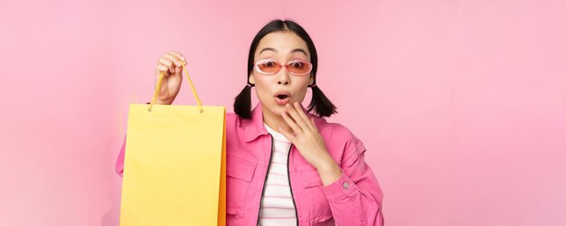 Einkaufen Stilvolles asiatisches Mädchen mit Sonnenbrille, das Tasche aus dem Geschäft zeigt und lächelnd Verkaufspromo im Geschäft empfiehlt, das über rosafarbenem Hintergrund steht