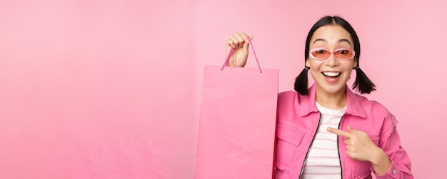 Einkaufen Stilvolles asiatisches Mädchen mit Sonnenbrille, das Tasche aus dem Geschäft zeigt und lächelnd Verkaufspromo im Geschäft empfiehlt, das über rosafarbenem Hintergrund steht