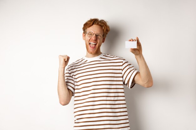 Einkaufs- und Finanzkonzept. Junger Mann, der Bankpreis gewinnt, Plastikkreditkarte zeigt und Faustpumpe macht, vor Freude und Zufriedenheit schreit, weißer Hintergrund