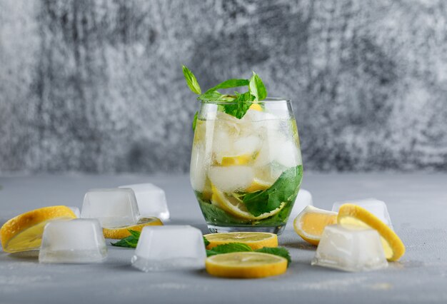 Eisiges Entgiftungswasser in einem Glas mit Seitenansicht von Zitrone und Minze auf grauer und Grunge-Oberfläche