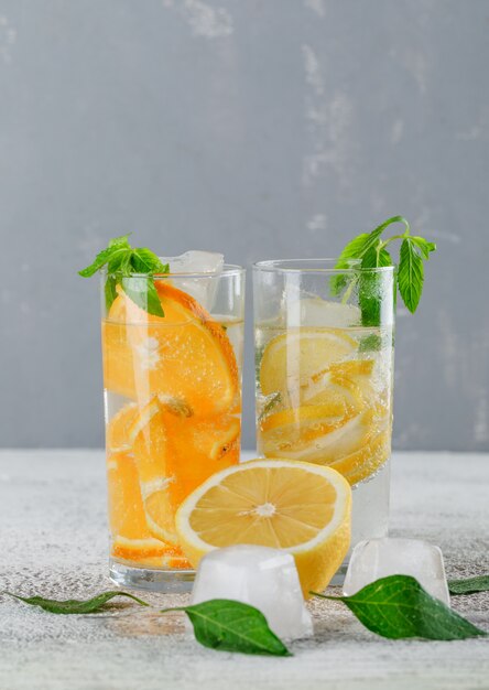 Eisiges Entgiftungswasser mit Orange, Zitrone, Minze im Glas auf Gips und Schmutzwand, Seitenansicht.