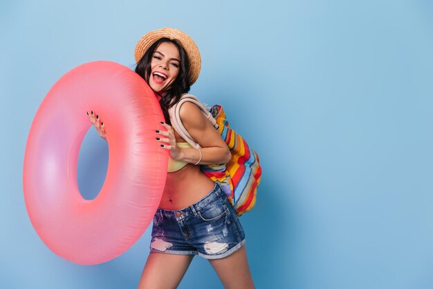 Ekstatische brünette Frau mit rosa Schwimmkreis Studioaufnahme eines anmutigen Mädchens mit Strohhut, das auf blauem Hintergrund tanzt