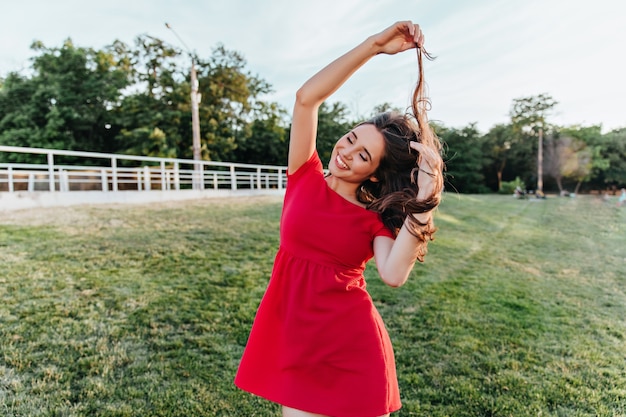 Ekstatische junge Dame in der Sommerkleidung, die mit ihren Haaren während des Fotoshootings im Park spielt. Außenaufnahme des niedlichen Mädchens im roten Kleid, das Spaß am Wochenende hat.