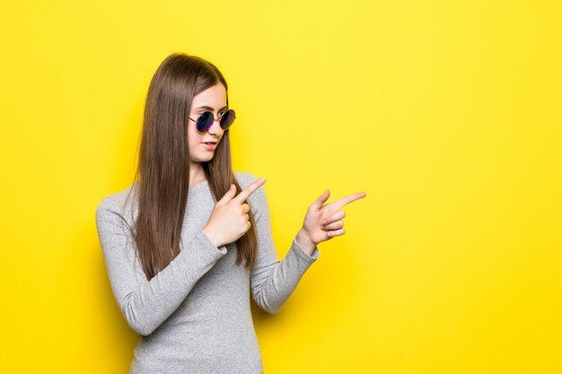 Ekstatische junge Frau in den Jeansshorts, im weißen Hemd und in der Sonnenbrille, die Hand auf Hüfte halten, wegschauen und zeigen