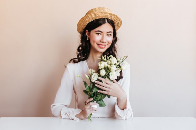 Ekstatische koreanische Frau, die beim Aufstellen mit Blumen lächelt. Blithesome lockige asiatische Frau, die weiße Eustomas hält.