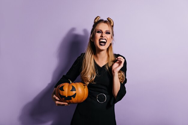 Ekstatische langhaarige Frau, die Halloween-Kürbis hält und lacht. Foto des emotionalen Vampirmädchens im schwarzen Kleid.