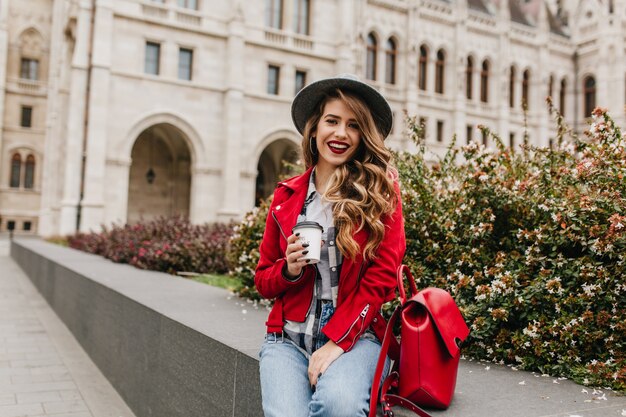 Ekstatische lockige Frau in der roten Jacke, die Kaffee vor dem schönen alten Gebäude trinkt