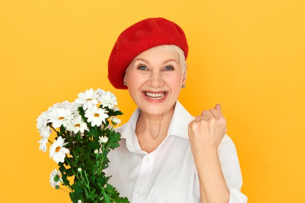 Ekstatische modische reife ältere Frau in der roten Haube, die einen Blumenstrauß hält