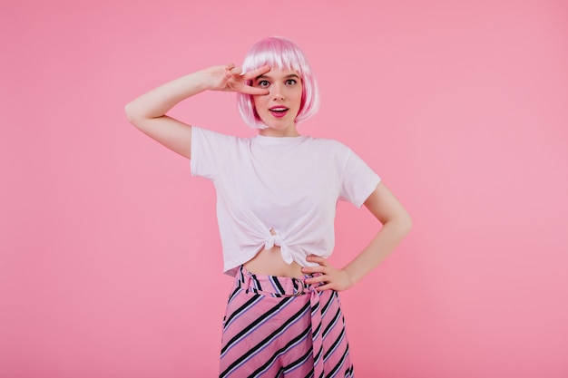 Ekstatisches formschönes Mädchen in der trendigen Kleidung, die mit lustigem Gesichtsausdruck auf rosa Wand aufwirft. sorgloses weibliches Model in weißem T-Shirt und glänzender Perücke