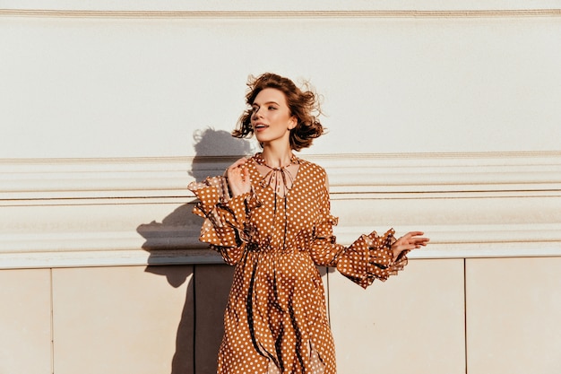 Ekstatisches kaukasisches Mädchen mit kurzen dunklen Haaren, die sich umsehen. Außenfoto der entzückenden weißen Frau im stilvollen braunen Kleid.