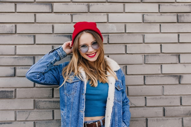 Ekstatisches kaukasisches Modell mit hellbraunem Haar, das mit Lächeln neben Backsteinmauer aufwirft. Außenporträt der sorglosen jungen Frau trägt trendige Jeansjacke.