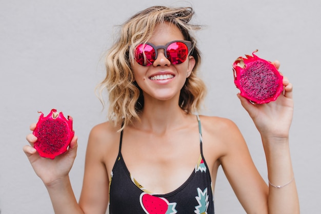 Ekstatisches lockiges Mädchen im schwarzen Kleid, das mit exotischen Früchten aufwirft. Innenporträt der inspirierten Dame mit roter Pitaya lokalisiert.