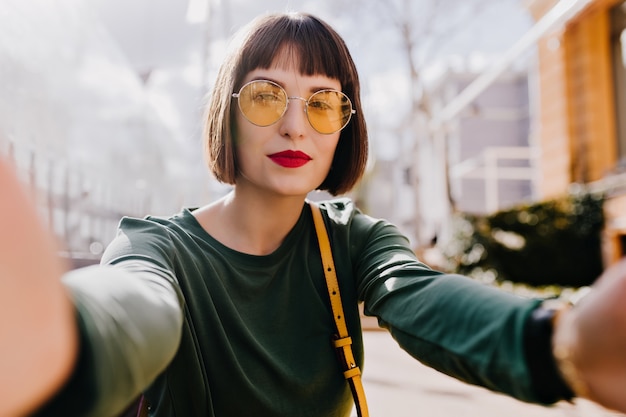 Ekstatisches Mädchen in der gelben Sonnenbrille, die selfie mit ernstem Gesichtsausdruck macht. Außenaufnahme der erfreuten brünetten Frau im grünen Pullover, der Bild auf Stadt macht.