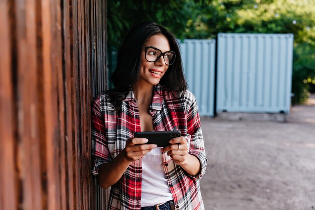 Ekstatisches weibliches Modell in der Brille unter Verwendung des mobilen Internets. Außenporträt des glamourösen Mädchens, das auf der Straße mit schwarzem Smartphone steht.