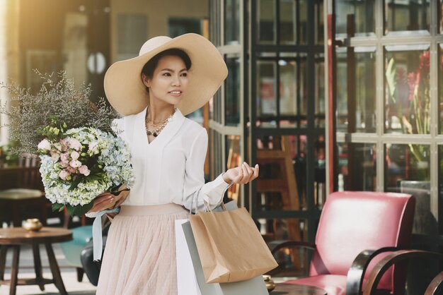 Elegante asiatische Dame, die aus Café mit Einkaufstaschen und Blumenblumenstrauß heraus geht