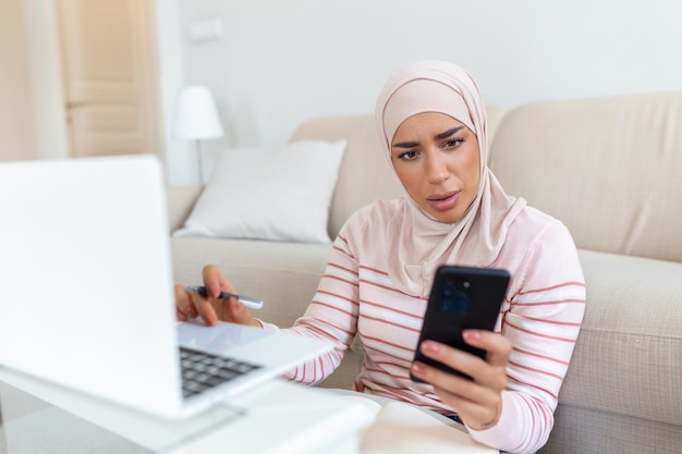 Elegante, attraktive muslimische Frau, die mit Mobiltelefon und Laptop Online-Shopping-Informationen im Wohnzimmer zu Hause durchsucht Porträt einer glücklichen Frau, die Produkte über Online-Shopping kauft