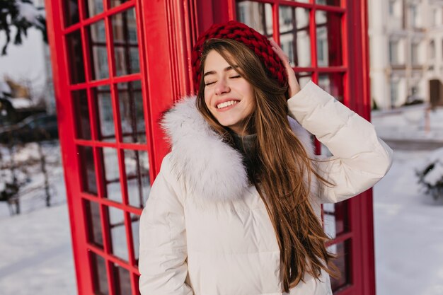 Elegante braunhaarige Frau, die mit romantischem Lächeln und geschlossenen Augen während des Winters in England aufwirft. Außenporträt der verträumten lächelnden Frau in der roten Wollmütze, die Fotoshooting nahe Anrufbox genießt.