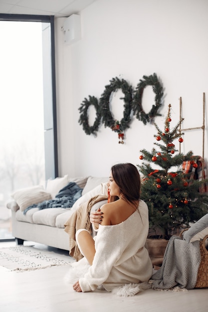 Elegante Dame nahe Weihnachtsbaum. Frau in einem Raum. Famale in einem weißen Strickpullover.