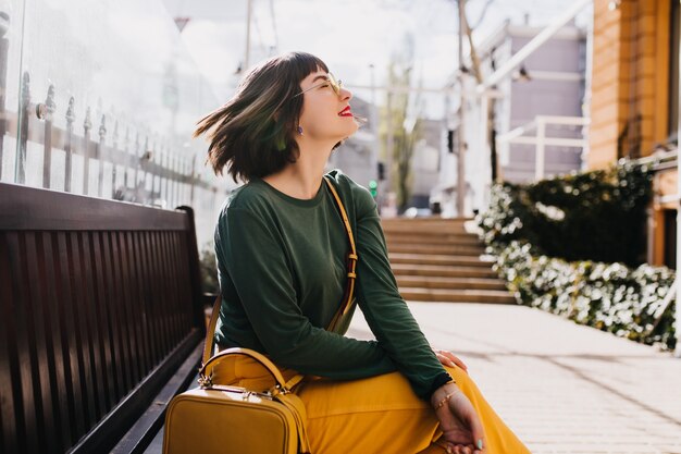 Elegante europäische Frau mit dem geraden kurzen Haar, das auf Bank sitzt. Außenporträt des erstaunlichen weißen Mädchens trägt grünen Pullover im Frühlingstag.