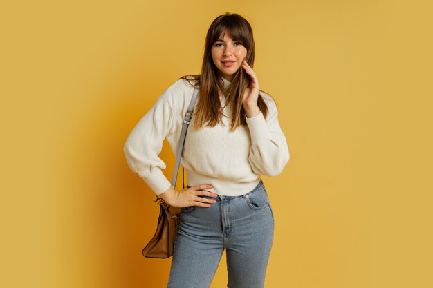 Elegante Frau, die im Studio auf Gelb aufwirft. Trägt einen weißen Wollpullover und Jeans.