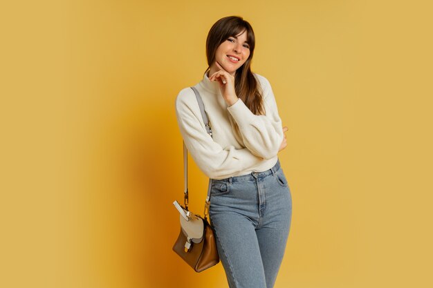 Elegante Frau, die im Studio auf Gelb aufwirft. Trägt einen weißen Wollpullover und Jeans.