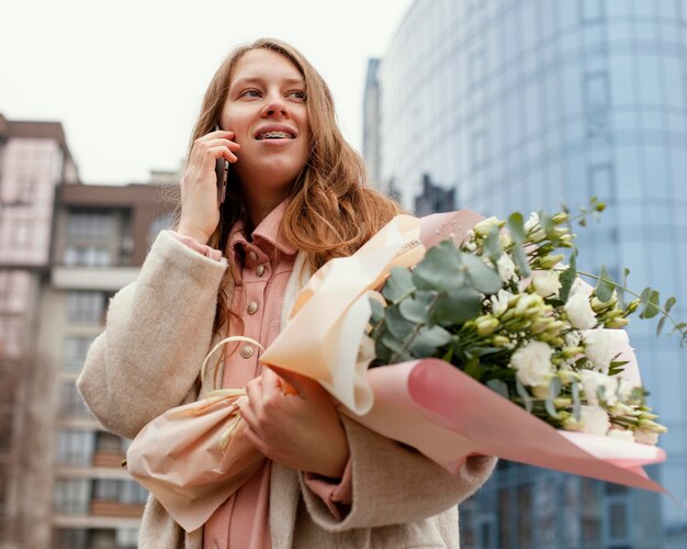Elegante Frau im Freien, die auf dem Smartphone spricht und Blumenstrauß hält